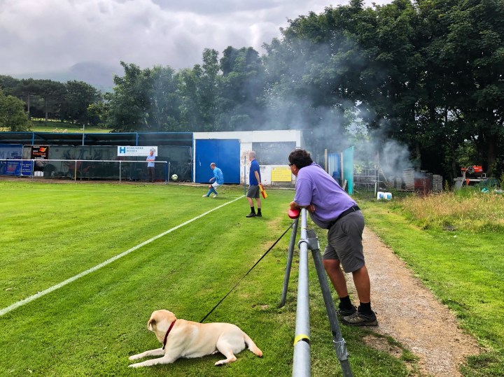 A man and his dog watch a Llandyrnog United player take a corner in a match against Penmaenmawr Phoenix, whilst smoke drifts over from a bonfire in the corner of the latter's Cae Sling ground