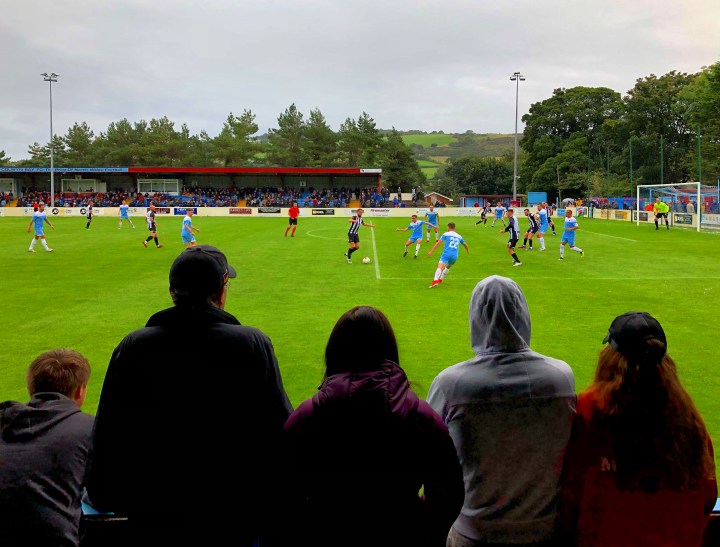 A group of five people watch Colwyn Bay and Llandudno play a match in front of a big crowd at Llanelian Road