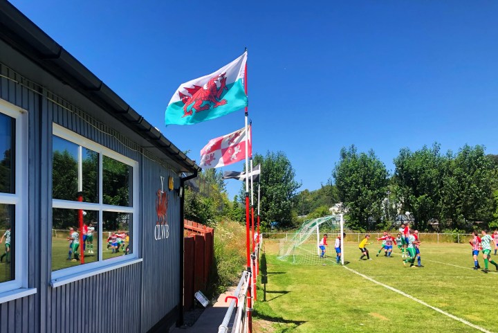 Players of Glan Conwy and Abergele compete in the air for a corner on a sunny afternoon at Glan Conwy's Cae Ffwt ground, with the action reflected in the clubhouse windows