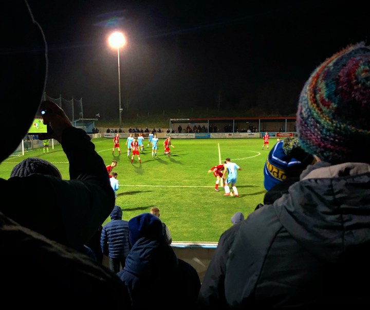 Spectators in hats and gloves watch, and one films on his phone, as Colwyn Bay attack during a night game against Buckley Town