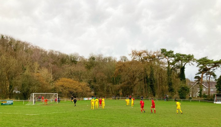 Spectators and players watch on as Y Felinehli's number seven gets ready to take a late penalty in front of a backdrop of brown trees