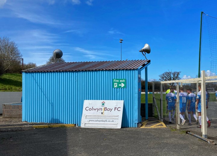 Colwyn Bay players return to the field from the tunnel; a disused fixtures board is propped up behind the small corner stand at Llanelian Road