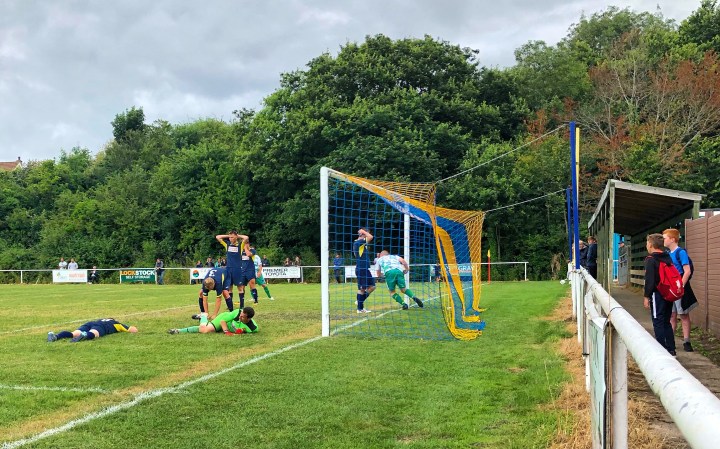 Two Llandudno Junction players stand with their hands on their heads, whilst a Mochdre Sports player gathers the ball from the back of the net after Sports had equalised in a match at The Flyover Ground