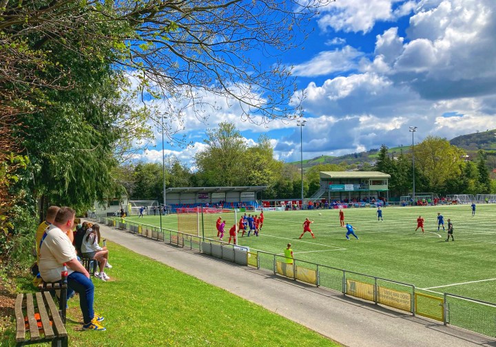 Fans watch from the grass bank at Newtown as Denbigh Town defend a corner against Trethomas Bluebirds 