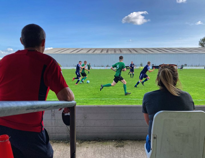 A man and a woman watch as players from Greenfield and Rhos Aelwyd run past them in front of the large corrugated metal building that sits next to Greenfield's pitch