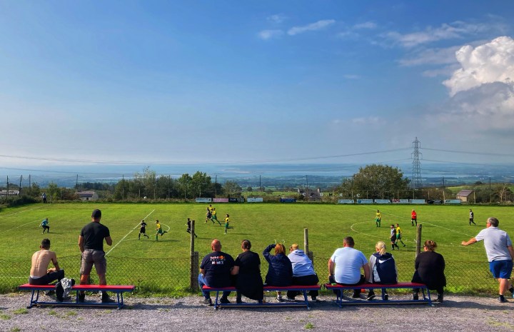 Supporters sit on a bench watching Mountain Rangers take on Gaerwen; the pitch is beneath the fans on a plateau, beyond the pitch the landscape stretches out across the Menai Straits to Ynys Mon