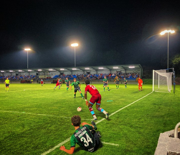 A prone Aberystwyth Town player watches on as Colwyn Bay's Udi Akpan shapes to cross during a night match at Bay's Llanelian Road ground