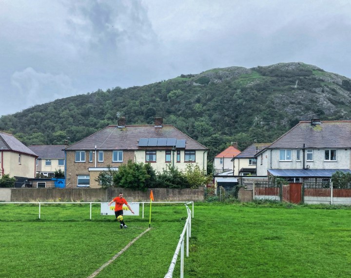 A Meliden player takes a corner kick in the rain in front of the houses that back onto his side's pitch