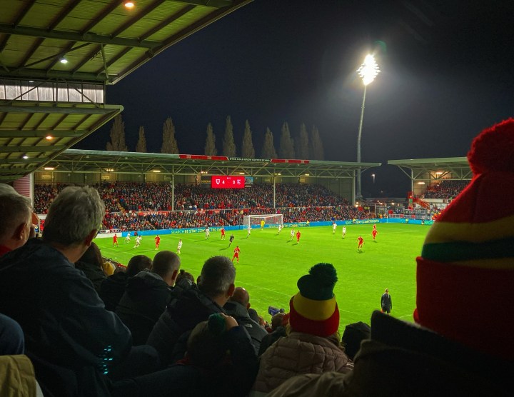 Wales fans, some in bobble hats, watch on as their team take on Gibraltar under the floodlights at the Racecourse Ground