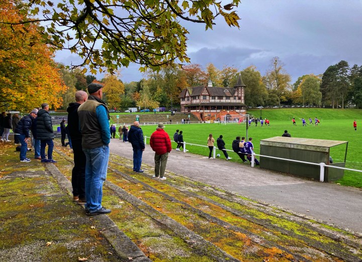 Spectators on a mossy terrace beneath autumnal trees watch a match in front of Cadbury Atheltic's ornate Victorian clubhouse