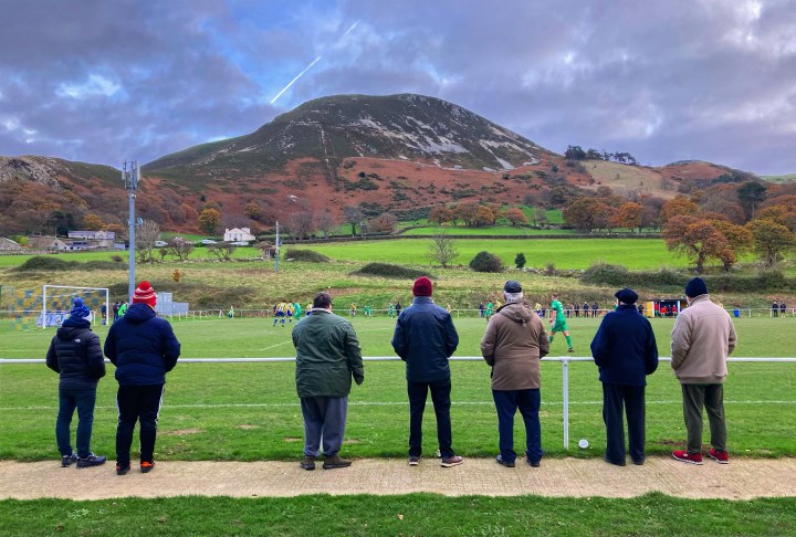 Seven older spectators stand and watch as Penmaenmawr Phoenix play Rhyl 1879 in front of the mountain at Cae Sling