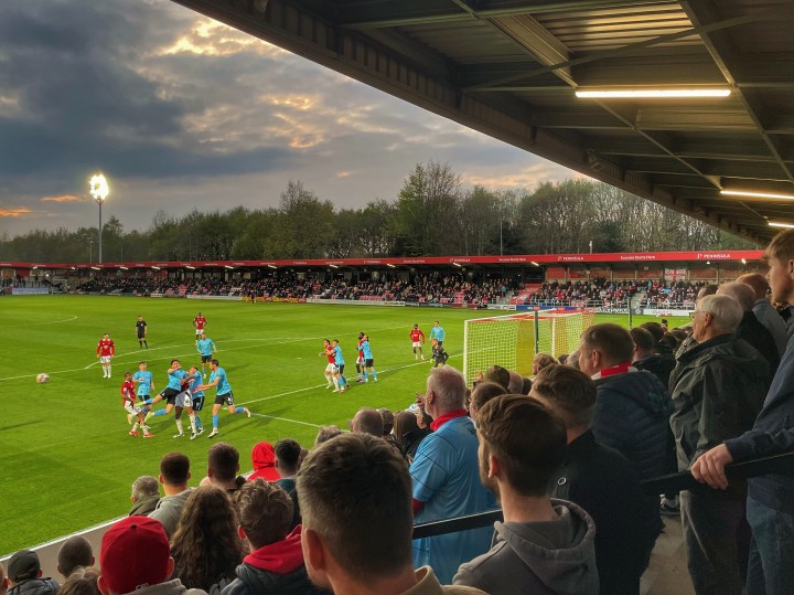 Doncaster fans watch on from the away terrace as their team defend a Salford City corner during an evening match at City's Peninsula Stadium
