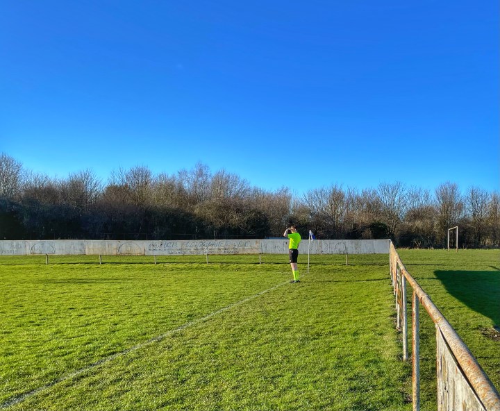 A lonely assistant referee shields his eyes from the sun in front of long faded advertising boards at Mold