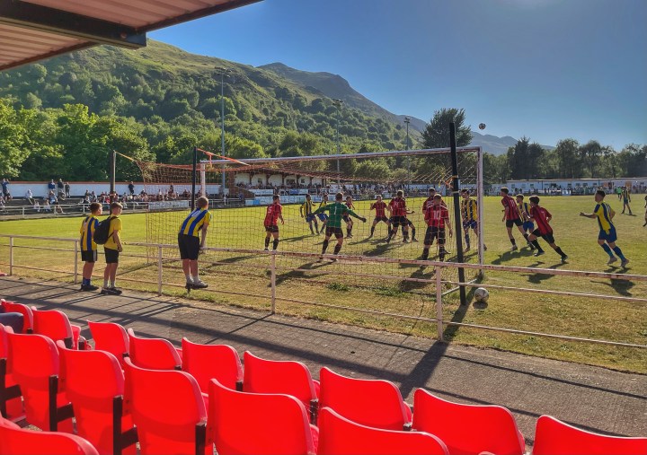 Three young fans in blue and yellow Penmaenmawr shirts watch on from the fence on a very sunny evening at Conwy Borough as Penmaenmawr Phoenix have an attacking corner against Llanfairfechan Town