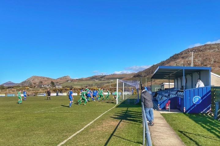Nefyn United players attack a corner in a home match against Boded; mountains and blue sky are visible beyond the pitch