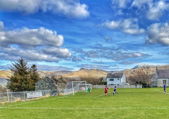 A Bontnewydd player volleys a shot wide at Mynydd Llandygai in front of a background of mountains topped with snow