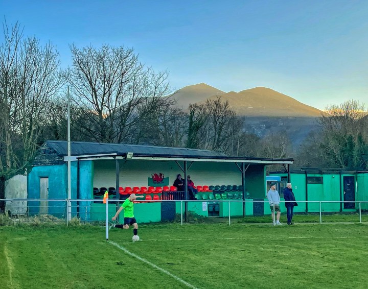 A Bethesda Athletic player takes a corner in front of a small stand; in the background the mountains catch the evening sun