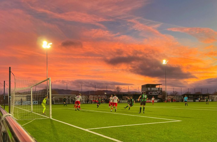 A Penmaenmawr Phoenix player takes a shot away against Llandudno Amateurs as the sun sets of the Maesdu Park stadium