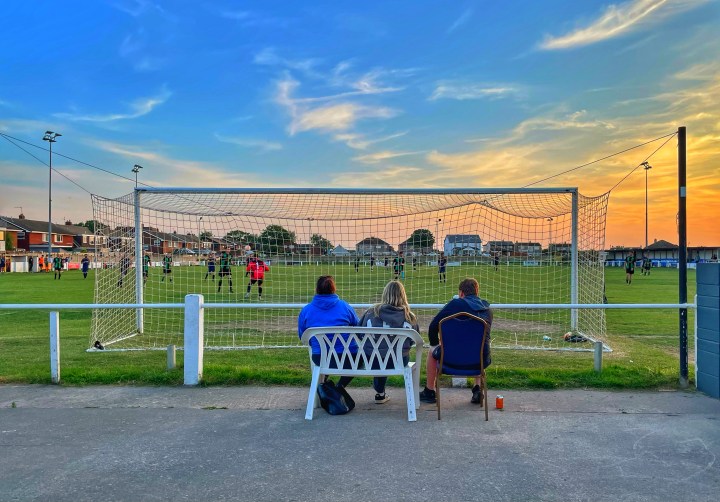 Three Penmaenmawr Phoenix fans, one on a dining chair, two on a plastic bench, watch on from behind the goal as their team play against Bow as the sun sets at Prestatyn