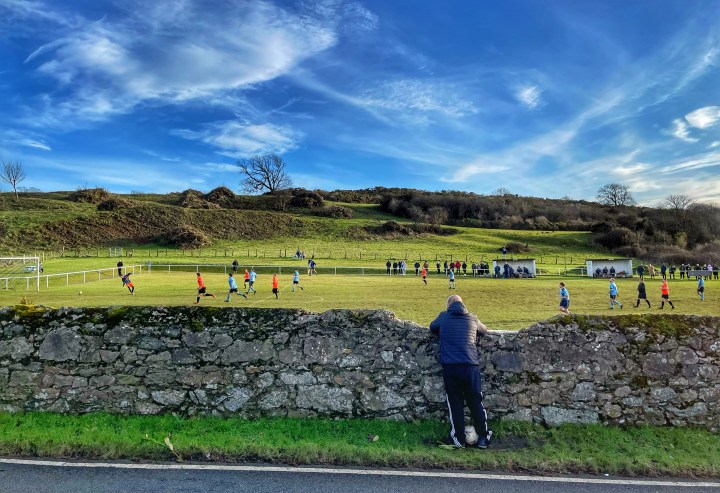 A man with a football at his feet watches from behind a stone wall as Pentraeth take on Penrhyndeudraeth