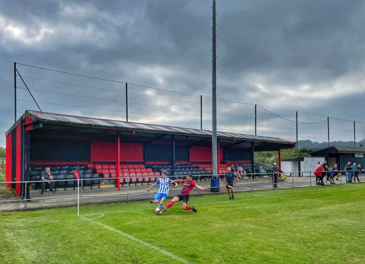 A Holyhead Hotspur winger is prevented from crossing the ball by a sliding tackle from a Porthmadog player in front of a near-empty stand