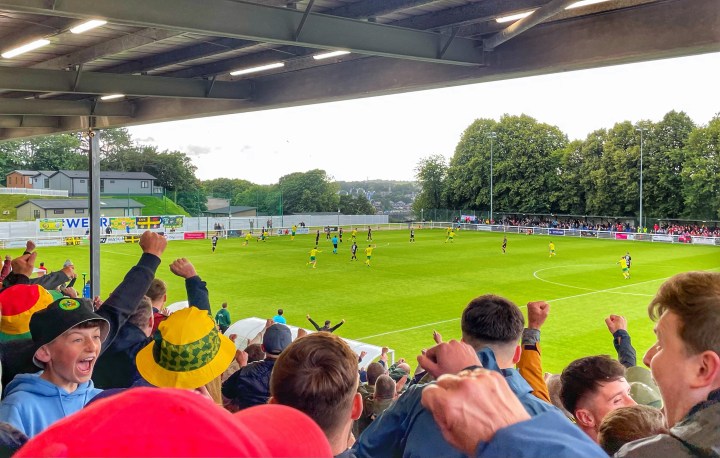 Caernarfon Town fans celebrate in the stands as behind them on the pitch their team go 1-0 up against Crusaders at Bangor's Nantporth Stadium