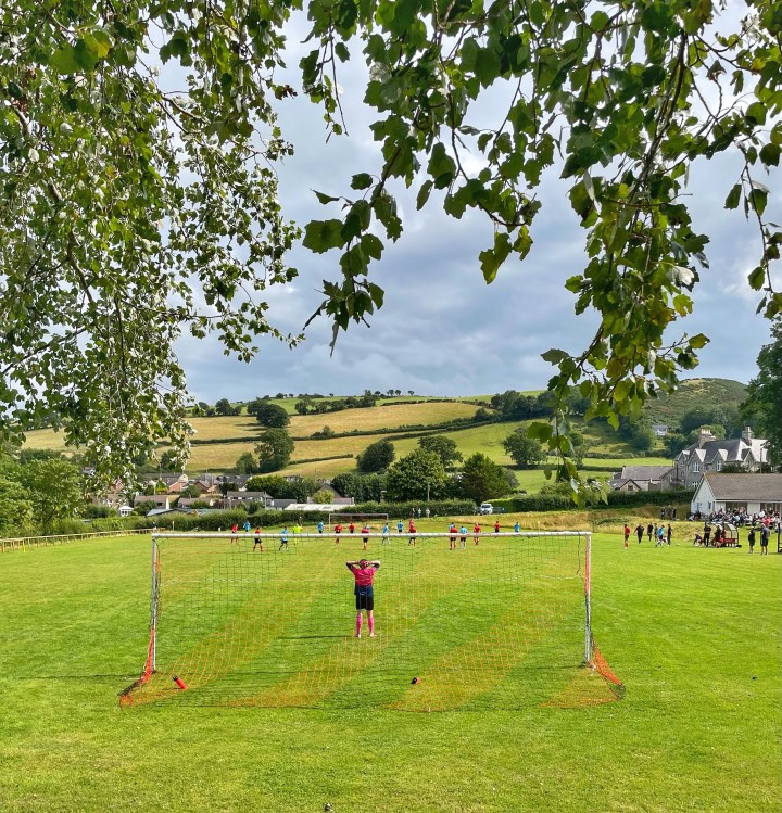 A football match in rolling country hills viewed from behind one goal. The match action is at the other end of the field. The goalkeeper at the near end stands with his hands on top of his head.
