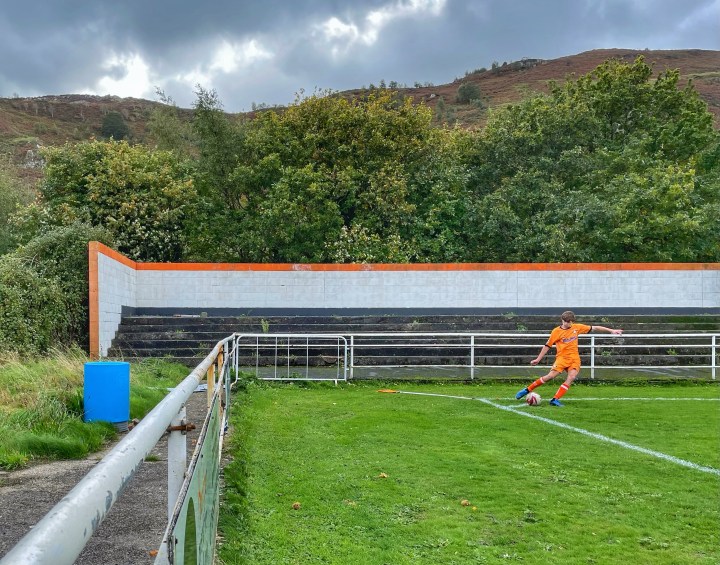 A Conwy player in their distinctive orange kit takes a corner in an otherwise empty corner of the ground; the flag has fallen over, mountains rise up behind him.