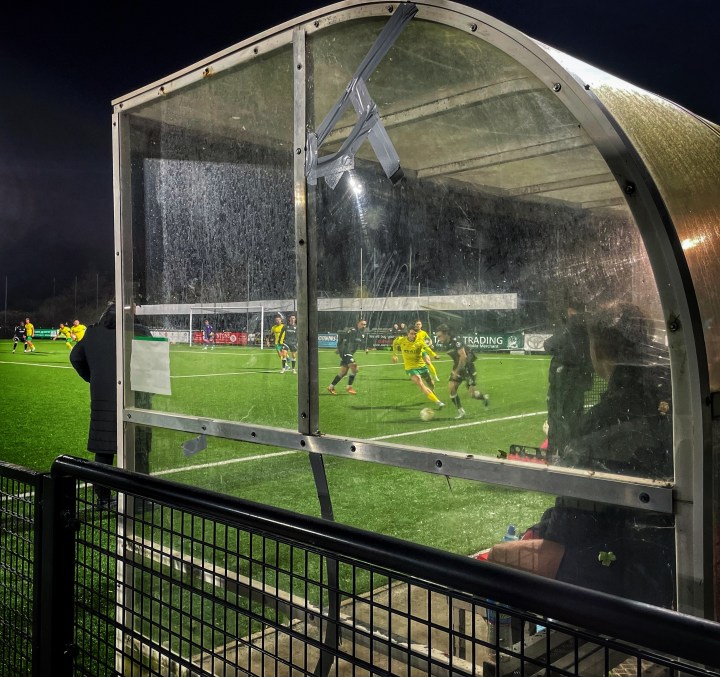 Night match action glimpsed through the side of a perspex dugout at Llandudno as Caernarfon play The New Saints.