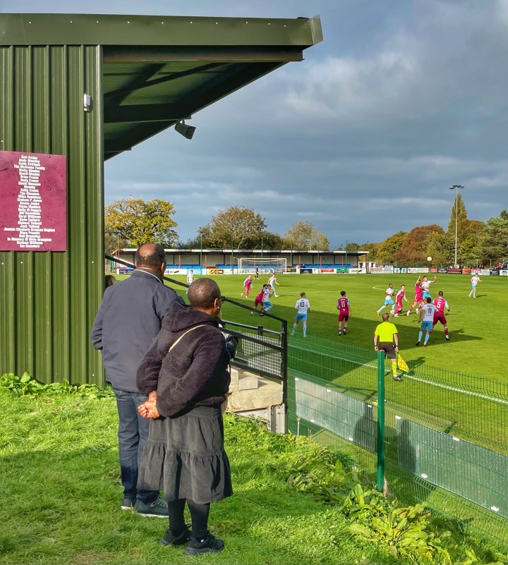 A middle aged couple watch Colwyn Bay take on Cardiff Met from a grass bank beside the main stand.