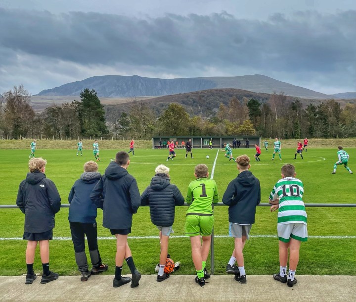 A group of young lads watch on from the fence as Talysarn Celts host Llanerch-y-Medd against a mountainous background.