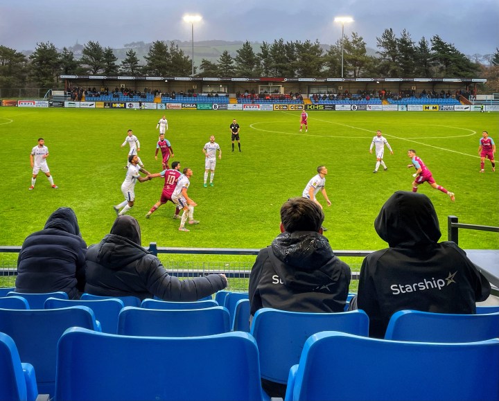 Fans watch on from the seats of the main stand as Colwyn Bay and Penybont players get ready to challenge for a header in heavy rain.