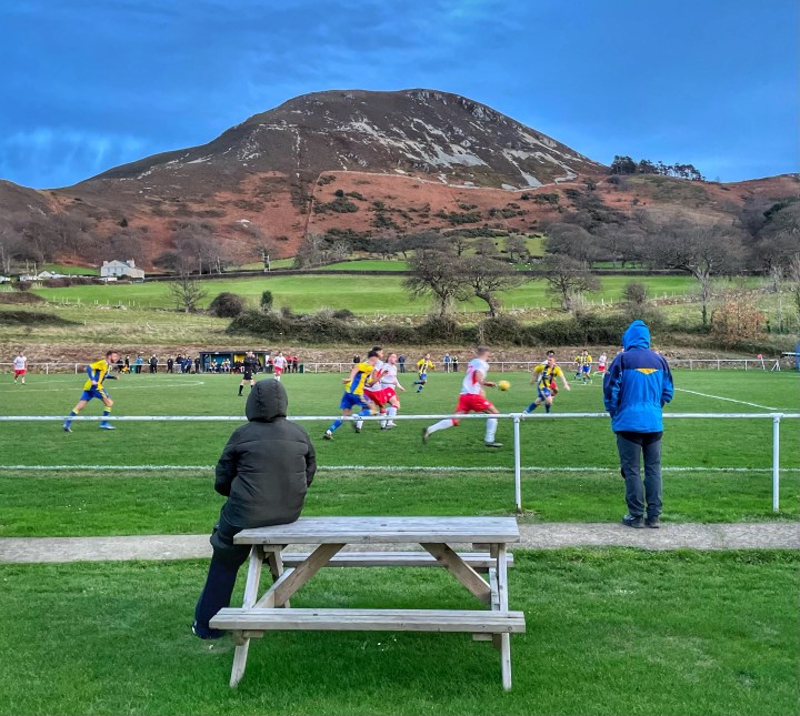 A fan sits on a picnic table and watches Penmaenmawr Phoenix play Mumbles Rangers against a mountainous backdrop.