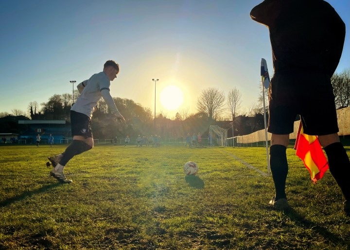 A Caersws player runs up to take a corner into a low sun at Mold Alexandra; beside him the assistant referee shields his eyes.