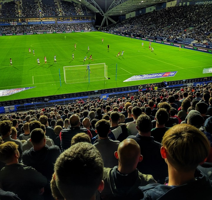 A night match at Huddersfield Town viewed from the back of the away end, thousands of heads illuminated by the floodlights watch a Doncaster Roves attack.