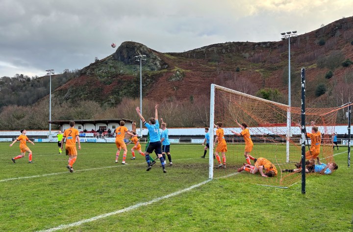 Llansannan players appeal to the referee for a goal as members of their own team and opponents lie tangled in the goal mouth and net at the latter team's mountain backed home ground.