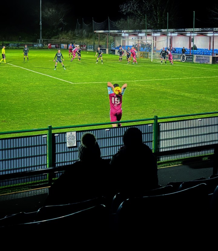 Two supporters watch on as Colwyn Bay's Matty Hill launches a long throw-in towards the penalty area during a night match against The New Saints.