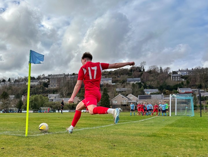 A Cemaes Bay player takes a corner during their 4-3 defeat to Penrhyndeudraeth; beyond the pitch the town of Penrhyndeudraeth climbs the hill side. 
