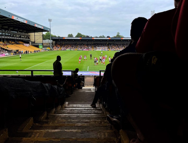 Match action during Port Vale vs Doncaster Rovers viewed from the shadowy depths of the stands on a sunny day; spectators are silhouetted against the sunshine.