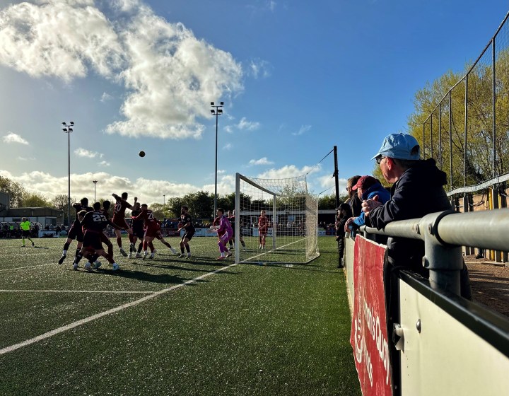 A Colwyn Bay fan in a sky blue gap watches his team tussle beneath a corner kick with hosts Connah's Quay Nomads against a low sun at Flint Town United.