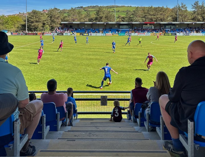 A very young Colwyn Bay fan sitting on the steps of the main stand watches his team take on Penybont in bright sunshine.