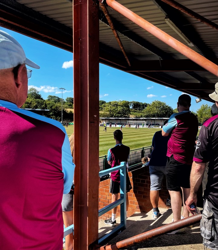 Colwyn Bay supporters in shorts and claret and blue Colwyn Bay shirts watch on from the stands. 
