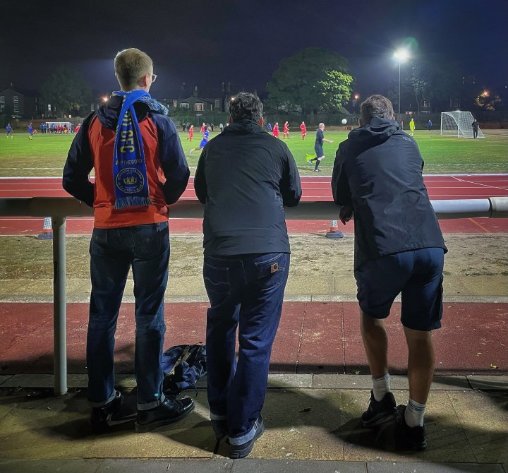 A trio of Lewisham Borough supporters watch their team during a night match from behind a railing and a long jump pit.